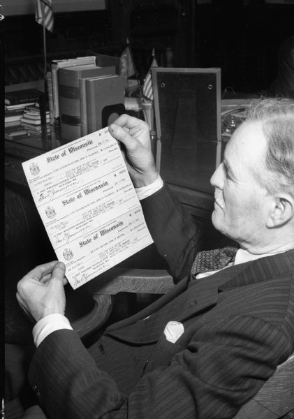 Fred Zimmerman, Wisconsin Secretary of State, holding three checks dated January 31, 1944.  The checks are payable to First Wisconsin National Bank, Milwaukee, for $15 million and $10 million; and First National Bank, Madison, for $1 million.