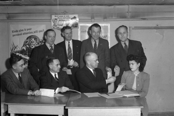 Seven members of the Madison Chapter of CUNA presenting checks for War Bonds to H.C. Jamieson, co-chair of the Dane County War Bond Committee, seated next to Jayne Eustance.  Standing left to right: Fred Mason, Henry Johnson, Ralph Runge, W.C. Tomkins; seated: John Williams, George Timmerich, H.C. Jamieson and Jayne Eustance.
