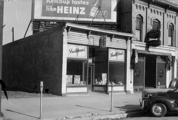 Exterior view from street towards the Oscar Fridblom's Health Food Store at a new location, 120 East Washington Avenue. A Heintz ketchup billboard is sitting on top of the store.