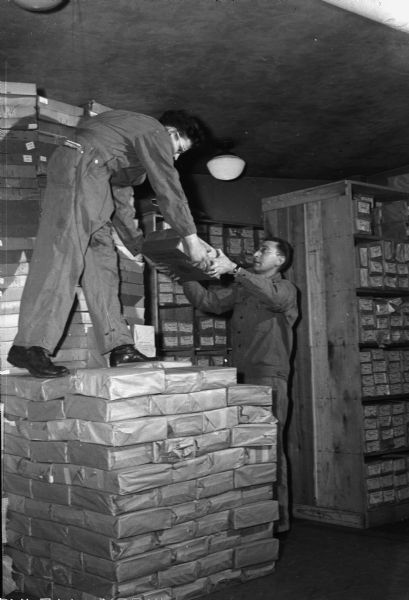 Corporal Harold Haven standing on a stack of packaged books, handing packages to Sargeant Russell West at the United States Armed Forces Insitute supply depot. The books are used by the United States Armed Forces Institute (USAFI), in lessons for soldiers and sailors at the fighting fronts of the world. The lessons are corrected by the University of Wisconsin - Extension.