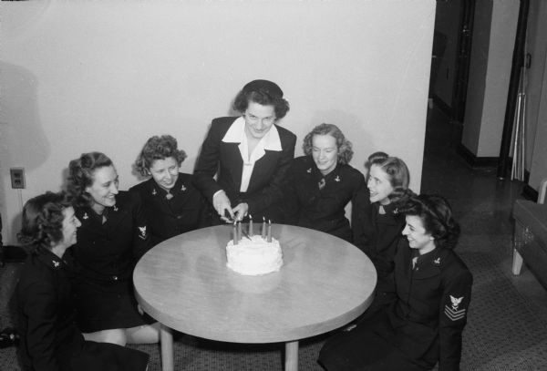 Slightly elevated view of group of women gathered around a table. "Leap year baby" of 1924, Anita Burr, is shown with a birthday cake, after enlisting in the WAVES (Women Accepted for Volunteer Emergency Service) station at the University of Wisconsin. Helping her celebrate, left to right: WAVES Kay Campbell, Lillian Skott, Mary Hagenah, Ruth Marcus, Norma Powell, and Angie Patrice, all in uniform. Miss Burr is in the center, cutting the cake.