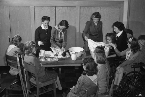 Dane County Unit of the Association for the Disabled training members of Girl Scout Troop No. 6 at the Washington Orthopedic School for Crippled Children, 545 West Dayton Street. The children are learning about the care of infant and small children. Left to right, facing the seated scouts are Mrs. Rufus Wells, instructor; Mary Parks, first class Scout; Erna Schweppe, troop leader and teacher at the school, and Winifred Orton, assistant troop leader, who is seated and has a Scout on her left.

