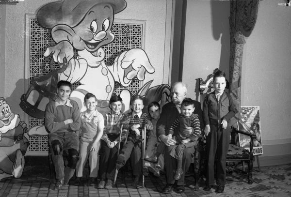 Governor Walter Goodland seated with handicapped children, probably in a movie theater. A large cutout of Dopey, from Snow White and the Seven Dwarfs, is on the wall behind the children.