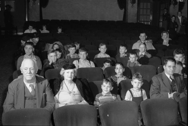 Parents and children, including Governor Goodland, seated in a movie theater to watch Snow White and the Seven Dwarfs.