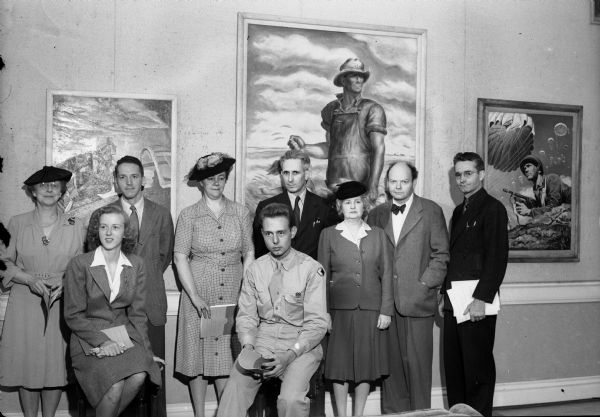 Nine people posed in front of "Our Good Earth" by John Steuart Curry at a Memorial Union exhibit of paintings executed for use on war posters.  Private Henning is seated, in uniform, Mrs. Walter Goodland (wife of Governor Goodland), is standing to his right, and John Steuart Curry is to her right. Lovey Pond is seated, Sarah Ross is on her left, and Mrs. Marshall Browne is on Lovey's right.