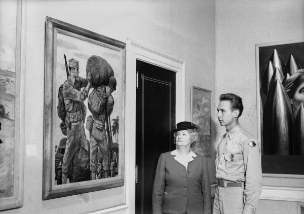 Mrs. Walter Goodland (wife of Governor Goodland), and Private Raymond J. Henning, former UW student, standing in front of "Carry Your Share!" by Joseph Hirsch at a Memorial Union exhibit of paintings executed for use on war posters.