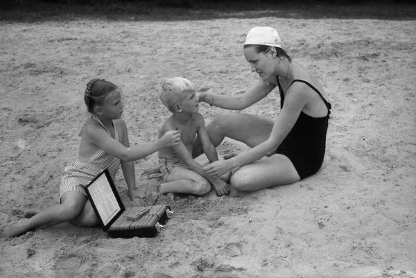 Linda Seed, lifeguard, is removing sand from the eye of John Hilhard with the help of Jackie Klopp, who is handing her an ampule of olive oil to soothe John's eye.