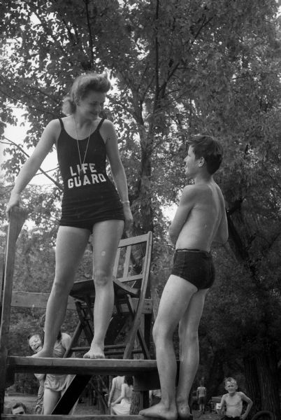 George Miller is reporting a problem to Lifeguard Lois Gardner. She is standing on a platform near a chair, and is wearing a whistle on a chain around her neck.