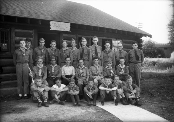 Outdoor group portrait of Boy Scout Troop 5, with Scoutmaster J.J. Feeney and Assistant Scoutmaster Bill Lavin. Photograph was taken in front of their new clubhouse constructed by Scouts' fathers.