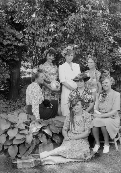 West Side Garden Club members are shown wearing hats made from plant materials. Standing in the rear are: Mrs. H.E. (Frieda) Consigny, Mrs. F.J. (Dagmar) Vea. and Mrs. Walter (Genevieve) Dakin. Mrs. F.D. (Laura) Chamberlain, sitting at extreme left, Mrs. (Myrtle) L.W. Ketchum, sitting center, Mrs. P.A. (Esther) Hauver, sitting far right, and Mrs. Herbert (Caroline) Kuentz, sitting on the ground.