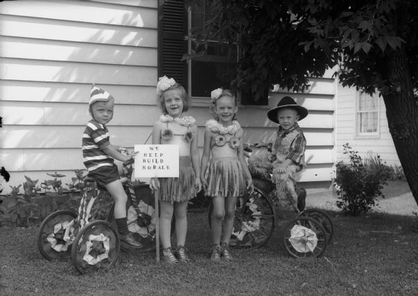 Children in costume for the Fourth of July parade in Westmorland neighborhood. From left to right are: Herman Heinecke Jr., son of Mr. and Mrs. Herman Heinecke, Joan Babcock, daughter of Mr. and Mrs. K.A. Babcock, Virginia Newbery, daughter of Mr. and Mrs. Raymond Newbery, and Scott Cameron, son of Mr. and Mrs. Frederick H. Cameron. They are shown with their sign: "We Help Build Morale."