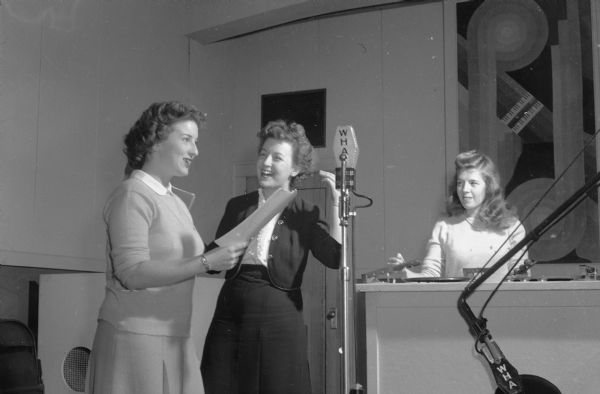 Three women at the summer radio workshop sponsored by WHA Radio on the University of Wisconsin-Madison campus. In the middle is Peg Bolger of the WHA staff coaching Jean Baillies at the microphone, while on the right Mary Anne Reed waits for her cue on the sound table.