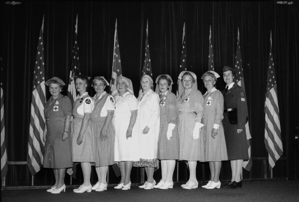 Nine Madison Woman's Club members posing in uniforms of their various Red Cross services. Left to right, Mrs. O.D.(Lillian) Smart, Mrs. William (Ivy) Fosdick, Mrs. Joseph (Salena) Wirka, Mrs. Ina Anderson, Mrs. Clara Flett, Mrs. William (Patricia) Werrell, Mrs. George (Mabel) Holdhusen, Mrs. Wilbur (Edith) Harris, and Mrs. (Sayda) Pettersen.