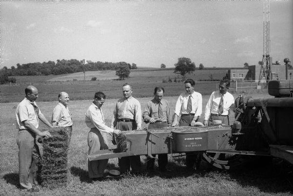 Six Russian representatives of the Soviet government observing a new McCormick-Deering Company Model 50-T hay baler at the Bowman Dairy Farm on Fish Hatchery Road. Left to right: Zakharchenko, Mayat, Kosilov, the leader of the group, Herasimov, Krasnichenko, and Ponstycin, with their interpreter, J. Shuldiner, second from left. In the background is a brick building and a large antenna.