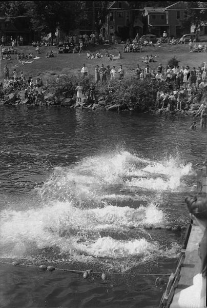 Annual city municipal swimming meet at B.B. Clarke Beach, with swimmers in the water, and observers along the shore, with cars, and houses in the neighborhood in the background.