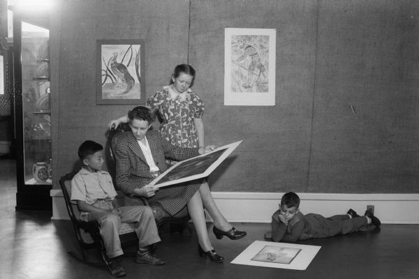 Miss Emilie Wiedenbeck, also known as Peter Mabie, writer and illustrator of children's books, is looking over some children's paintings and drawings with two fifth grade students, Billy Lee and Jacqueline Thompson. Michael Stein, a second grade student, is lying on the floor looking at a drawing. The students attend Washington School, and the art work will be on display at the State Historical Society museum in Madison.