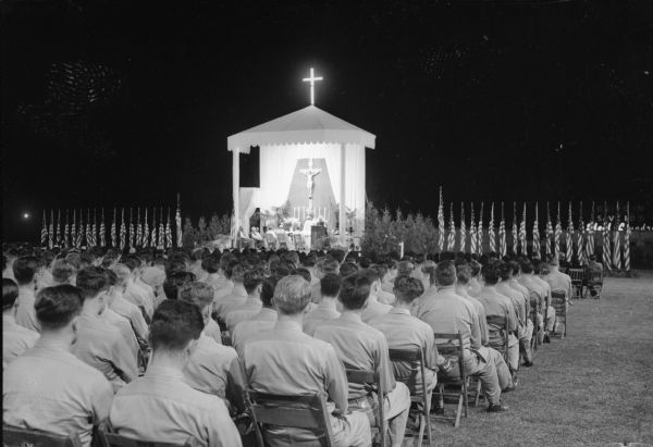 Third annual Holy Hour at Breese Stevens Field sponsored by the Holy Hour Society of Dane County, with flag flanked altar. Soldiers are sitting in the foreground. Rev. William P. O'Connor presided at the service. Nine-hundred Truax Field soldiers were present, having marched to the service from Truax Field.