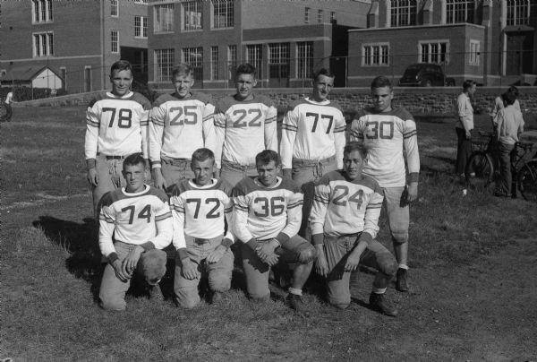Group portrait of nine East High School football players in uniform with the high school in the background. Standing from left: 78 Bob Kelly, 25 Bob Olson, 22 Duane Sydow, 77 Dick Lampe, 30 Jerry Wrend; kneeling from left: 74 Jim Toepfer, 72 Koltes, 36 Arnie Meier, 24 Homer Runkell.