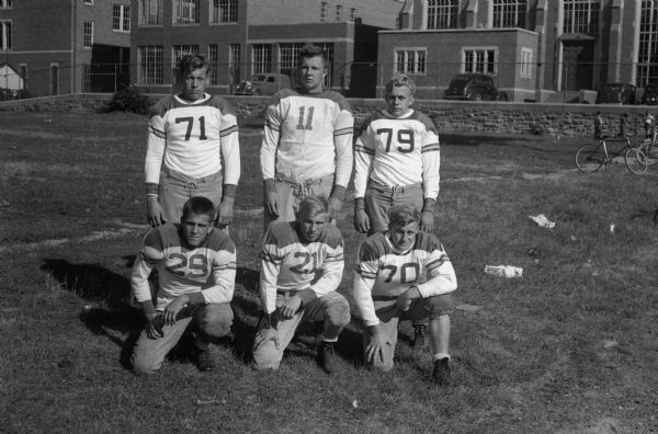 Group portrait of six East High School football players in uniform with the high school in the background. Standing from the left: 71 Stan Tjugum, 11 Leroy Anderson, 79 unidentified; kneeling from the left: 29 Bob Somerville, 21 Hanson, 70 Don Stevens.