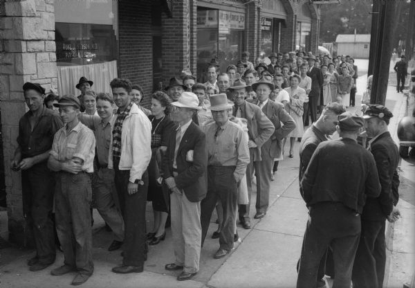 Slightly elevated view of customers standing in line on the sidewalk waiting to purchase shotgun shells at Berg Sporting Goods Store, 2123 Atwood Avenue. Other storefronts pictured include: 2121 Atwood, Dr. Robert J. Hudson, M.D.; 2119 Atwood, Modern Furniture Shop; 2117 Atwood, Richard Roman, optometrist and 2115 Atwood, Dutch Maid Ice Cream Shop.