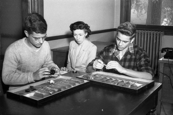Two students, Stanley Stemp Jr., Central High School, and Victor Brockmiller Jr., West High School, assemble mechanical devices as part of the vocational and social diagnostic testing offered by the Madison school system. Mrs. Marion Powers Topping, the assistant supervisor of the Board of Education, observes.
