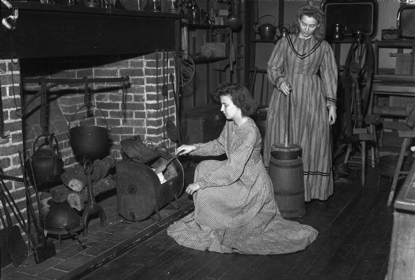 Joyce Elmer is basting a chicken in an iron reflector oven and Gene Lyte is using a butter churn. Both women are University of Wisconsin home economics majors who donned clothes and tried out the cooking facilities in the colonial kitchen in the Historical Society Museum on the 4th Floor of the State Historical Society Building.