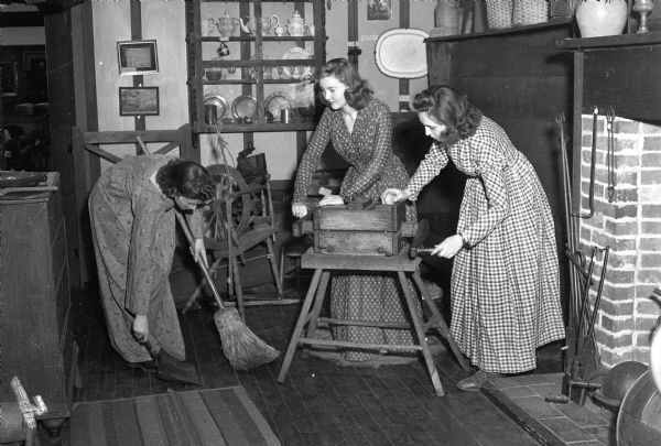Isomel Billings is using an old-fashioned broom, Lietzel Pelican and Patricia Cirves are operating a sausage grinder. The women are University of Wisconsin home economics majors who donned clothes and tried out the cooking facilities in the colonial kitchen in the Historical Society Museum on the 4th Floor of the State Historical Society Building