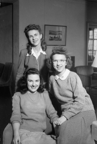 Three University of Wisconsin coeds of Elizabeth Waters Hall dormitory who have arranged for a tea and open house for facility members and friends. Standing is: Lucille McKeough Moser, Manitowoc, chairman of invitations and music; seated are Mary Jane Case, Ft. Atkinson, social chairman of the Hall; and Jeanette Kepke Thomas, Dodgeville, president of the dormitory.