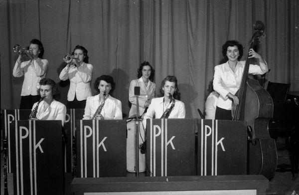 Group portrait of Peggy King's all-girl orchestra, posing with their instruments. The swing band plays on the weekends at fraternity and sorority parties. Left to right, front row: Iola Gage, Phyllis Trione, Maureen Millard, and Jewel Lubin.  Back row: Virginia Johnson, Peggy King, and Marjorie Johnson. Miss King is paying her way through medical school by heading the group.