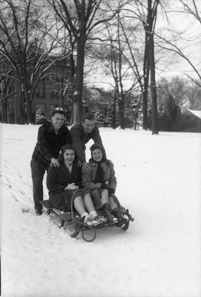 Four students sledding on Bascom Hill using a sled from 1838 which they borrowed from the Wisconsin Historical Society Museum. Seated from left: Joan Hammerstrom, Milwaukee, and Tommy Richey, Aruba, West Indies. Standing behind the sled from left: Al Tormey, Jr., Madison, and Larz Johnson, Appleton. The sled had been used near Sheboygan Falls for hauling supplies.