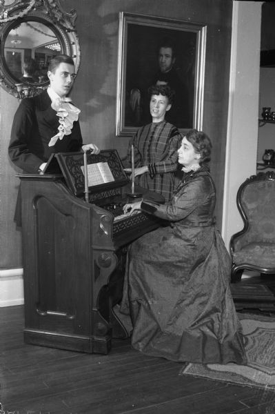 Reenacting a pioneer Christmas custom of many foreign settlers in the early days of Wisconsin of singing carols around the family organ, are left to right: Roy Vogelman, announcer at WHA, University of Wisconsin radio station; Mrs. Esther Hotten, announcer and engineer (seated at the State Historical Society Museum's old organ); and Mrs. Aline Hazard, director of the station's Homemaker's program. The group is dressed in costumes of the pioneer era. The museum's exhibition is called "Christmas Customs in Wisconsin."