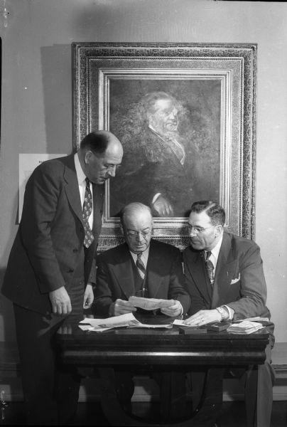 Members of the Wisconsin Banking Commission shown examining a State Historical Society collection of coins and currency, and bank notes signed by some of Wisconsin's greatest public figures, all dating back to territorial days. From left are E.W. Tamm, commissiion secretary: James B. Mulva, commission chairman: and H.A. Quinn, vice-chairman in charge of banks. The portrait in the background is of Alexander Mitchell, a famous early Wisconsin banker, financier and railroad executive.