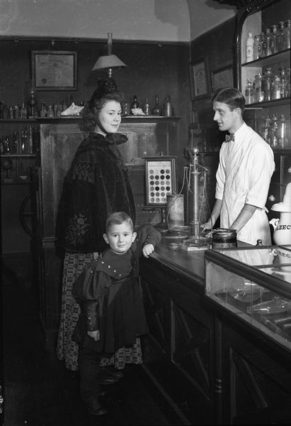 A woman, her son, and a sales clerk, all in period costumes in the drug store / pharmacy display in the Wisconsin Historical Society museum, then located on the fourth floor of the headquarters building.