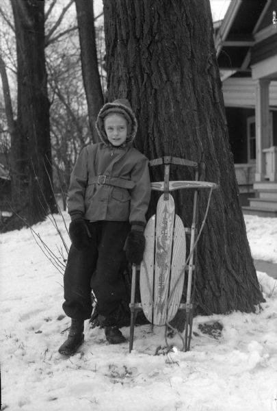 Winter scene with a young girl (possibly Polly Pyre, the daughter of Russell B. Pyre) in a snowsuit standing with an  "Airline Pilot" sled.
