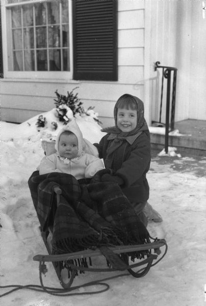 Sydney Ann, age 4, kneeling beside her baby sister, Stephanie, who is bundled into a sled.  The girls are the children of Mr. & Mrs. John Warren Fish, 816 Farwell Drive, Maple Bluff.