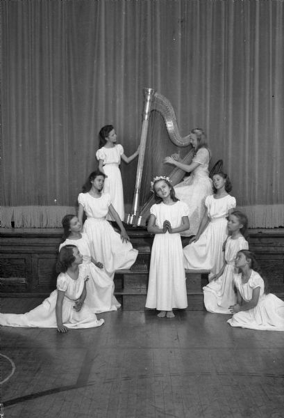 Eight girls (on the left side from floor to harp) Ruth Quale, Mary Fess, Patricia Kinney, Elaine Tripalin, (girl in the center) Carlene Stoker, (right side sitting on the floor Mary Alice Quale, next to her going up toward harp, Jean LeFebvre and Ruth Sund. The harpist is Patricia Runstrom.  They are at Lowell School for a Christmas party presentation titled "Ave Maria."