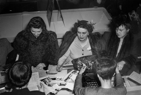 Three women buying war bonds at Manchester's Department Store while two clerks ring up the sale. The three women are all wearing fur coats, as it is a special Christmas holiday event.