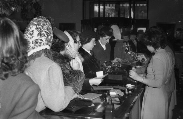 Women lined up along a sales counter at Manchester's Department Store to buy war bonds, while two clerks behind the counter ring up the sales.