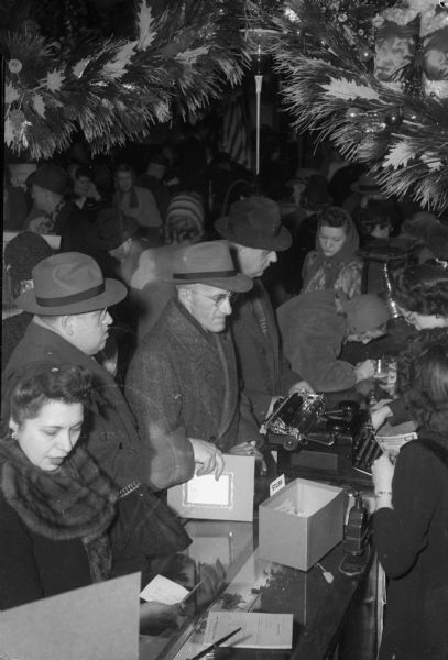 Men and women lined up along a sales counter at Manchester's Department Store to buy war bonds, while two clerks behind the counter are ringing up the sales.