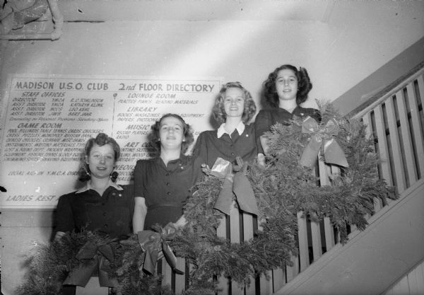 Senior Service Girl Scouts of Troop 22 from East High School posing on the stairs after having hung Christmas greens at the USO club. The girls are (left to right) Delores Stark, Rita Jambura, Eleanor Lunde, and Marian Jambura. A sign in the background lists the services of the USO club.