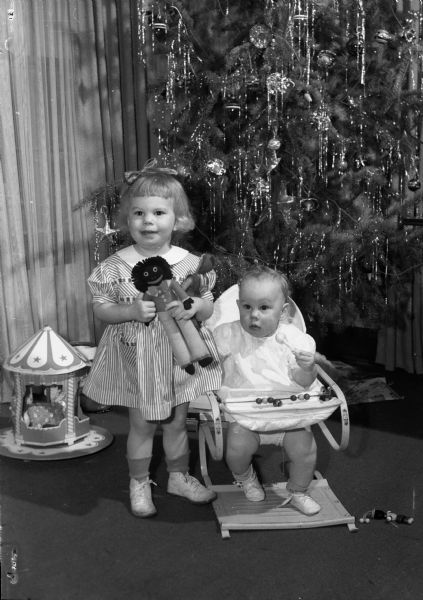 Cynthia and Catherine Rogers, daughters of Captain and Mrs. (Virginia) Sion C. Rogers, in their home celebrating Christmas. Cynthia, two-years-old, is holding two dolls, Catherine, ten-months-old, is seated in a jumper chair.  The dolls were made by their father when he was stationed in Australia.