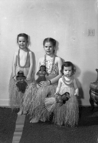 Karen, age 5, Gretchen, age 11, and Kristen, 16 months, daughters of Lt. and Mrs. Eldred Olson with their Christmas presents from Hawaii. They are wearing grass skirts, leis, and holding hulu dolls.