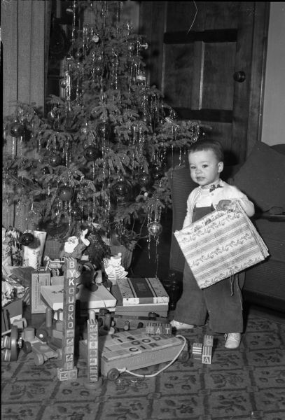Patricia Fitzpatrick, seventeen-months-old, holding a package and standing in front of the Christmas tree with presents surrounding her. She is the daughter of Lawrence and Frances Fitzpatrick. He is the managing editor of the <i>Wisconsin State Journal</i>.