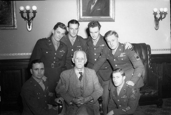 Governor Walter S. Goodland seated with six service men surrounding him. The men are promoting increased war production because of rationing of ammunition on the front lines of the military forces, by speaking to Badger Ordinance employees.
