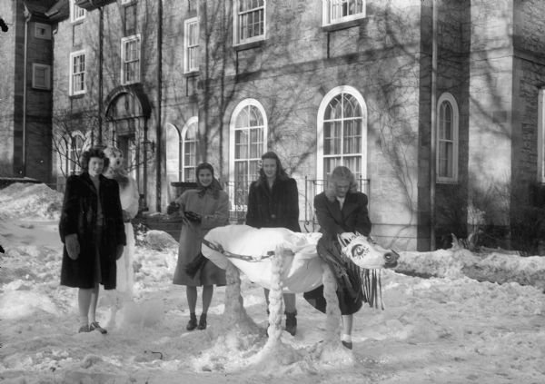 Winter scene with four young women, probably University of Wisconsin students, standing with a cow snow sculpture.