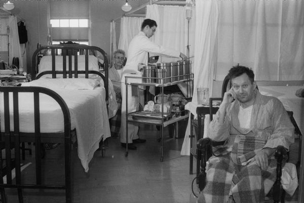 Wisconsin General Hospital room with two patients and a medical aide in crowded conditions.