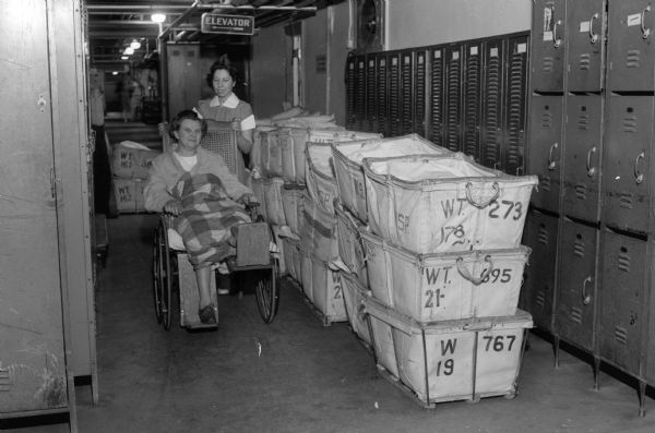 Patient being wheeled past laundry baskets and lockers in overcrowded Wisconsin General Hospital basement hallway.