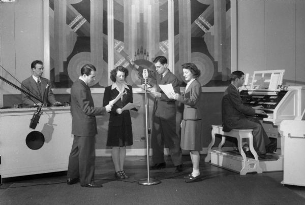 WHA studio, with two women and two men posing around a microphone. The woman to the immediate left of the microphone has been identified as Dorothy Shinstine, who worked in the offices of WHA.  In the background are (probably) Dan Frediani, the sound effects man, and Don Voegeli at the organ.