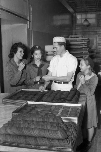 Three Girl Scouts sampling cookies at the Strand Baking Company, 2007 Atwood Avenue, with a baker, Reider Strand. Left to right: Sandra Bird, 1325 Jenifer Street, a member of Troop 40, East High School; Ruth Steinle, 211 North Dickinson Street, Troop 92, Lapham School; Mr. Strand; and Roberta Williams, 2321 Upham Street, Troop 73, Emerson School.