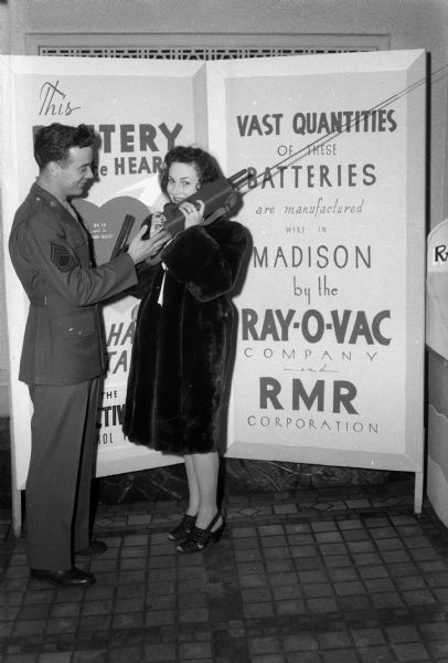 Soldier helping a woman use a Handie Talkie at the Ray-O-Vac & RMR promotional exhibit set up at the Capitol Theatre. "This battery is the heart of the Handie Talkie seen in the Warner hit 'Objective Burma.'"
